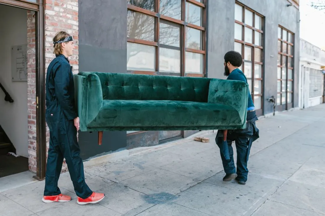 Zwei Umzugshelfer tragen ein Sofa über den Bürgersteig zum Wagen